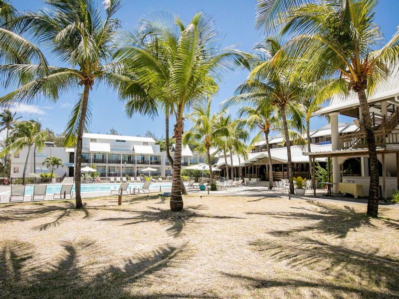 Hotel s bazénem, obklopený palmami a písčitým povrchem pod jasně modrou oblohou.