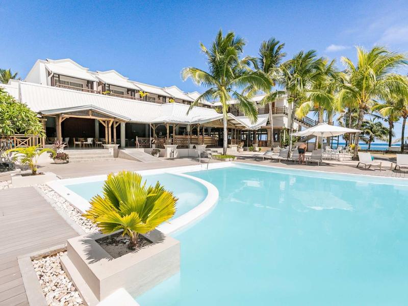 Hotel s velkým venkovním bazénem obklopený lehátky a palmami pod modrou oblohou.