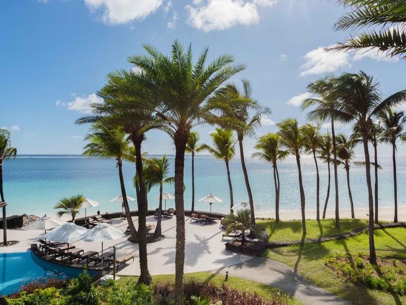 Palmy obklopují bazén s výhledem na bílou písečnou pláž a modré moře.