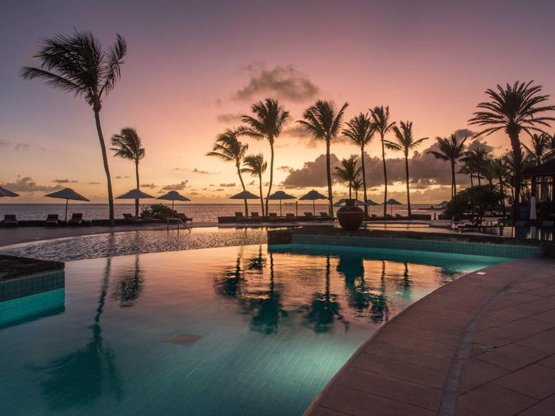 Luxusní hotel s palmami a velkým bazénem pod modrou oblohou.