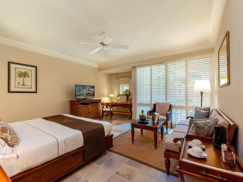 Světly hotelový pokoj s manželskou postelí, posezením, psacím stolem a velkým oknem s závěsy.