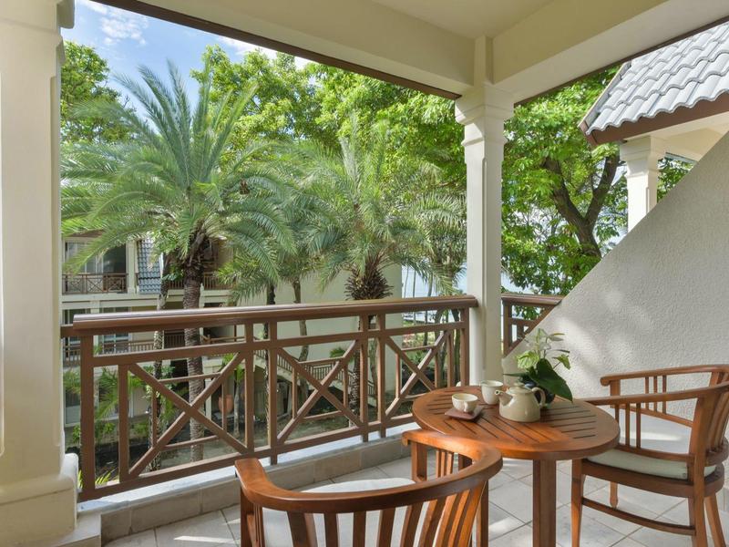 Balkon s dřevěným stolem a židlemi s výhledem na palmy a zelené listí.