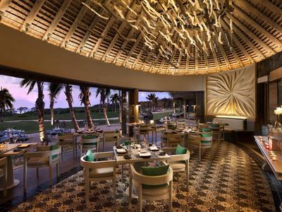 Elegantní hotelová restaurace s zakřiveným stropem, moderním osvětlením a výhledem na palmy za soumraku.