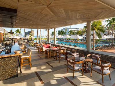 Zastřešená hotelová terasa s stolem, židlemi a výhledem na bazén a palmy.