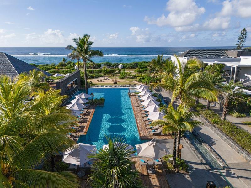 Pohled na bazén s lehátky, palmami a mořem v pozadí za slunečného nebe.