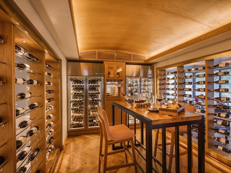 Elegantní vinný sklep s dřevěnými regály, skleněnými dveřmi chladničky a vysokým stolem se židlemi.