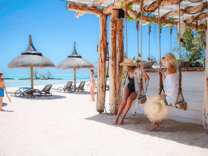 Strandscène met mensen op schommels onder houten pergola bij parasols en ligstoelen.