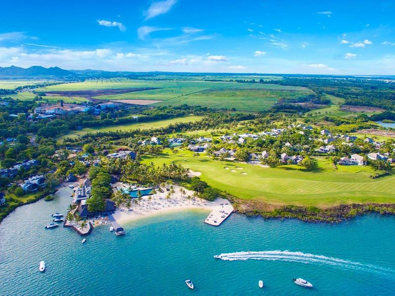 Kuststad met strand, boten op helder blauw water en groene velden onder een deels bewolkte hemel.