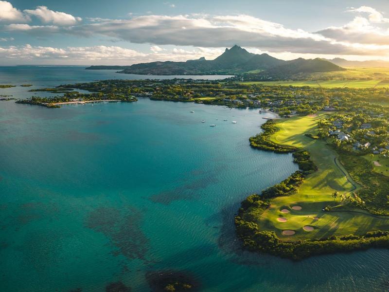 Luchtfoto van een kustlandschap met groene velden, rotsachtige kusten en een berg onder een deels bewolkte hemel.