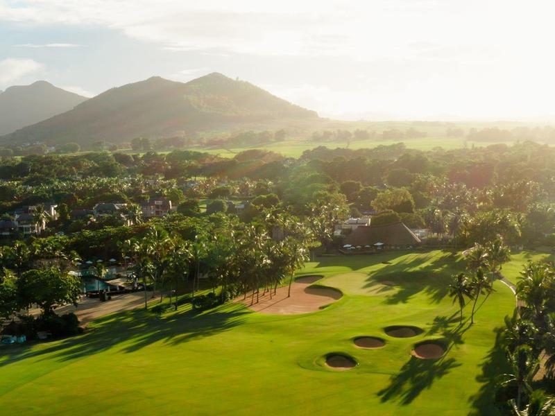 Zonnige golfbaan met weelderig groen en bergen in de verte onder een gedeeltelijk bewolkte hemel.