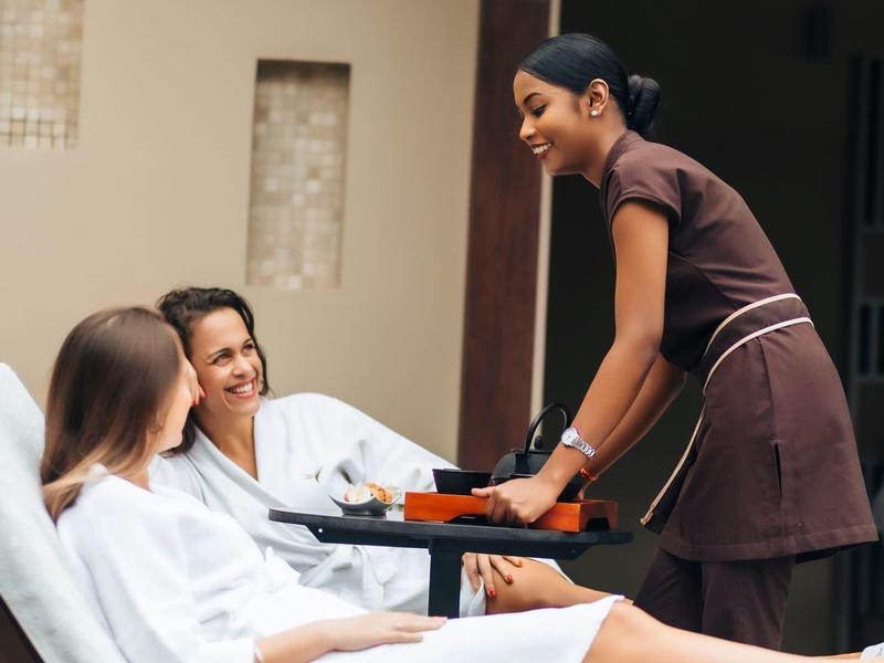 Twee gasten ontspannen in badjassen terwijl een glimlachende serveerster drankjes op een dienblad aanbiedt buiten.