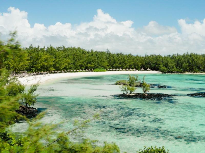 Wit zandstrand met turquoise water en groene bomen onder een blauwe lucht met wolken.