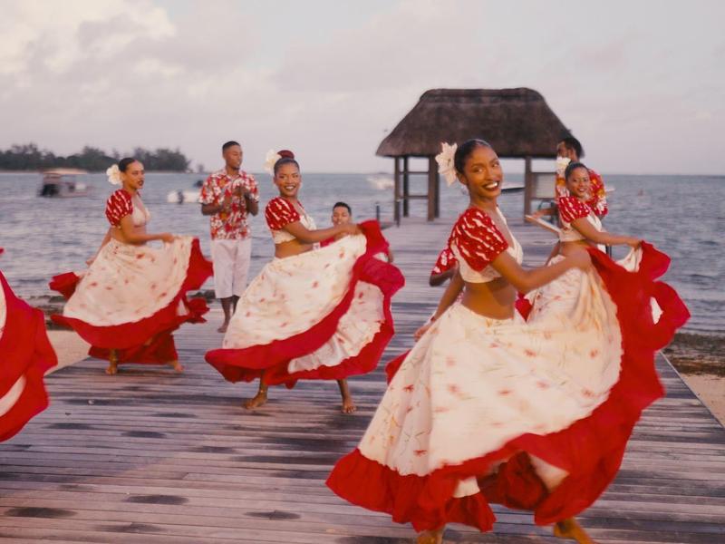Vrouwen in rood-witte jurken dansen bij het water met een houten prieel op de achtergrond.