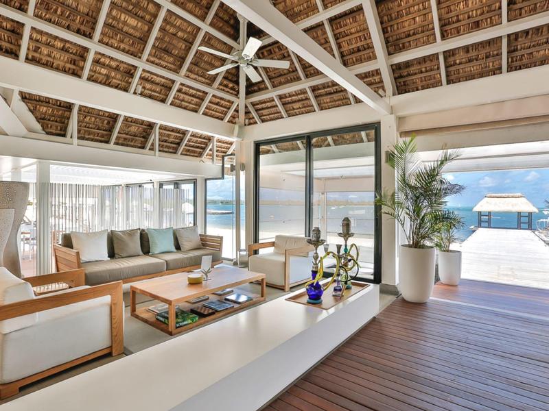 Lichte open lounge met houten meubels onder rieten dak met uitzicht op oceaan en zwembad.