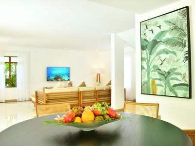 Soggiorno luminoso con divano in legno, grande quadro di piante e ciotola di frutta su tavolo rotondo.