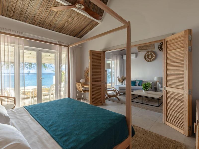 Chambre avec lit en bois et vue sur terrasse et mer à travers de grandes fenêtres.