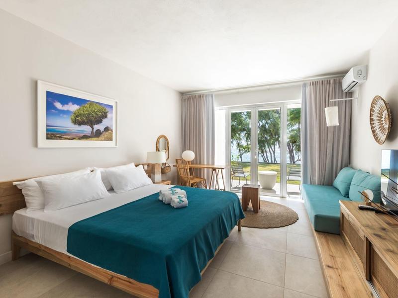 Chambre d'hôtel moderne avec lit double, meubles en bois, canapé et vue sur des arbres par grandes fenêtres.