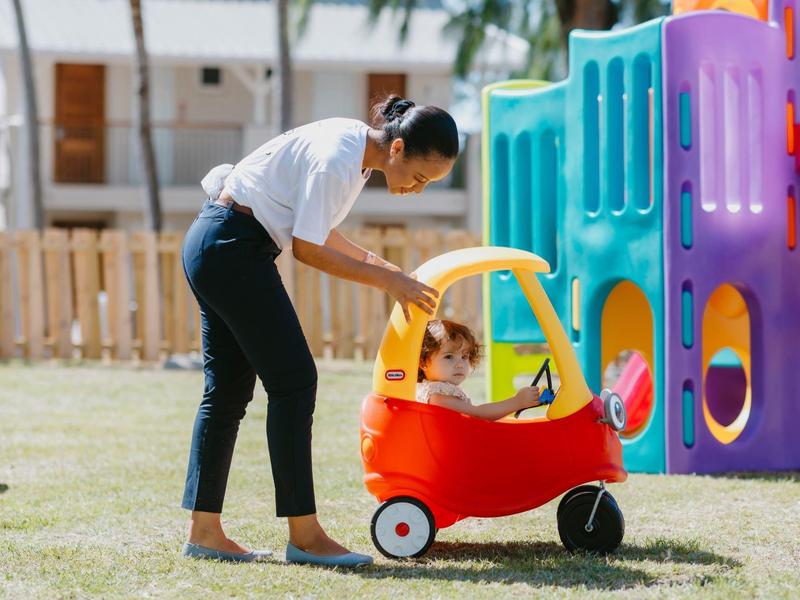 Femme jouant avec un enfant dans une voiture-jouet rouge devant une aire de jeux colorée