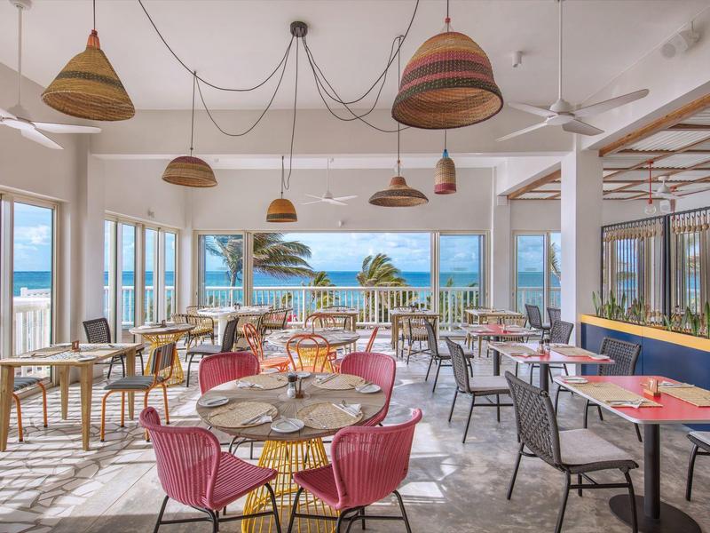 Licht, modern restaurant met kleurrijke stoelen en uitzicht op zee door grote ramen.