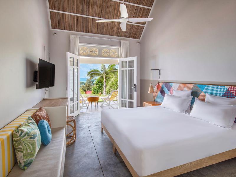 Lichte hotelkamer met tweepersoonsbed, kleurrijke wanddecoratie en uitzicht op terras met palmbomen.