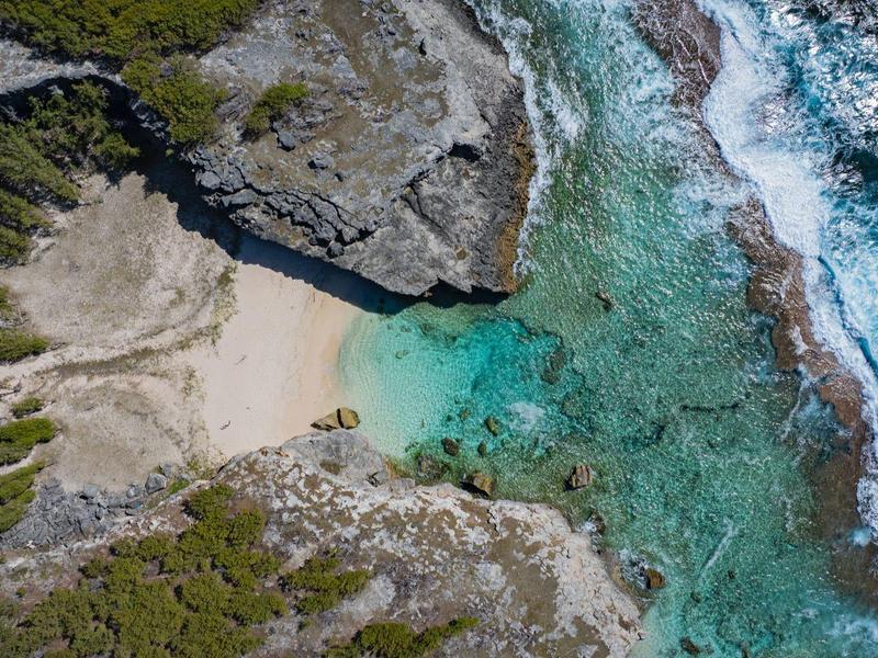 Luchtfoto van een afgelegen strand met helder turquoise water, omlijst door rotsen en begroeiing.