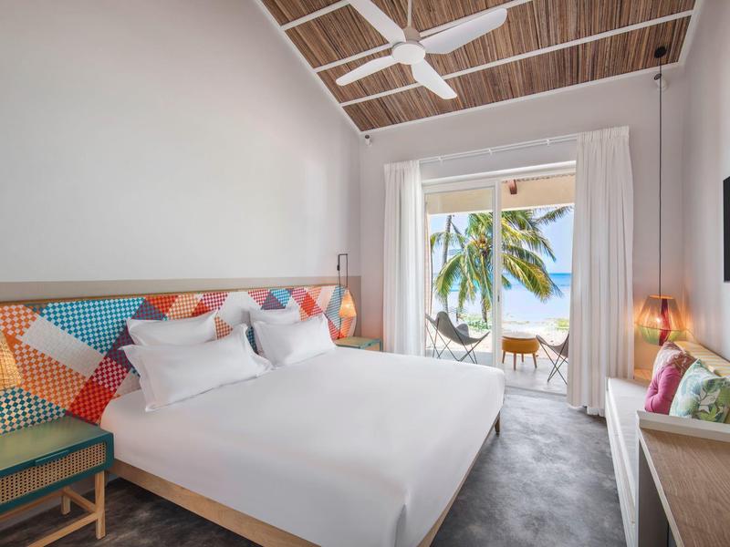 Lichte hotelkamer met plafondventilator, kleurrijk hoofdeinde en balkon met zeezicht.