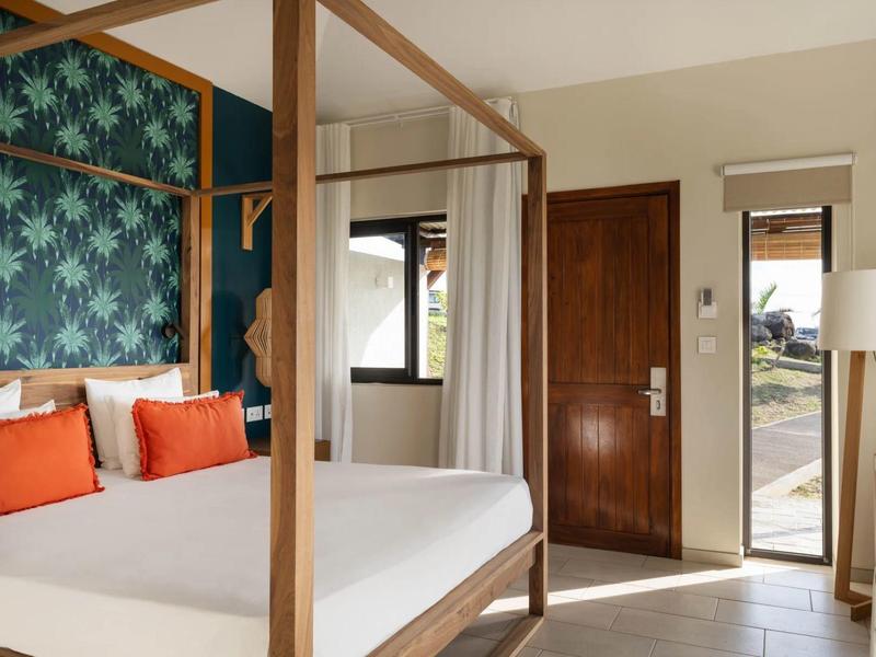 Světelný hotelový pokoj s baldachýnovou postelí, barevnými polštáři, přírodním dřevem a výhledem na venkovní dveře.