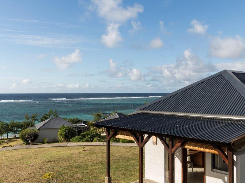 Výhled na verandu s dřevěnými sloupy a stoly, trávník a moře v pozadí.