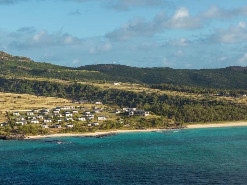 Pláž s modrým mořem a domy na úpatí zelených kopců pod modrou oblohou