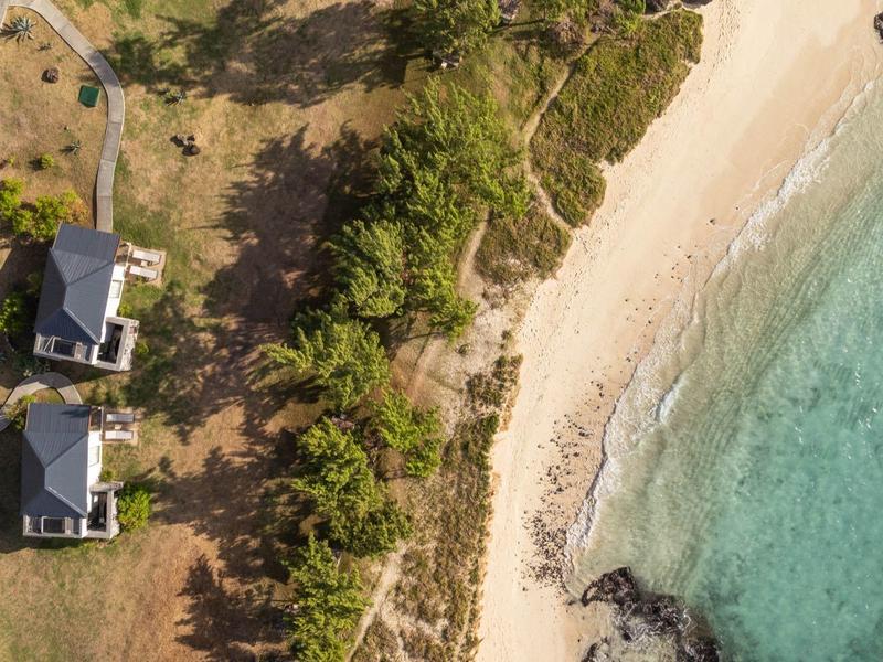 Letecký snímek dvou domů vedle písečné pláže s čistou modrou vodou a vegetací.