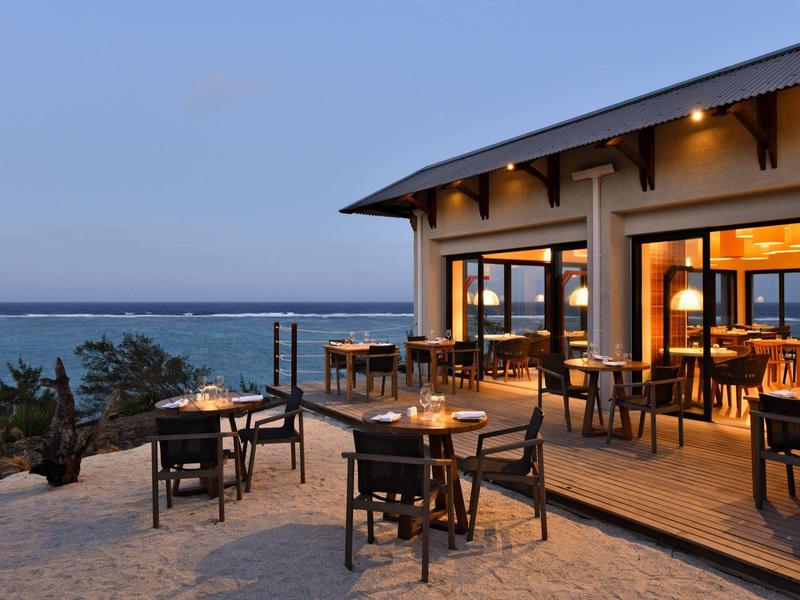 Venkovní restaurace s útulnými stolky na terase a v písku s výhledem na moře za soumraku.