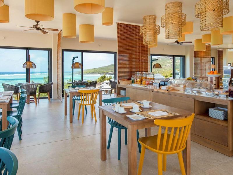 Světlá restaurace s dřevěným stropem, barevnými židlemi a výhledem na moře přes velká okna.