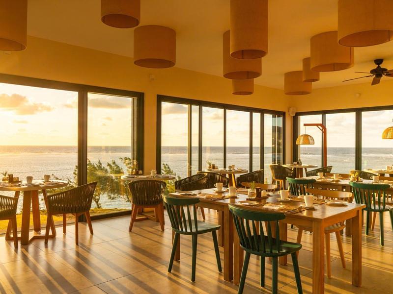 Světla restaurace s dřevěnou podlahou a velkými okny s výhledem na moře při západu slunce.