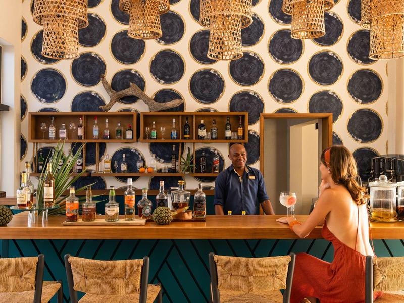 Elegantní bar s barmanem, ženou v červených šatech a pletenými koši visícími ze stropu.