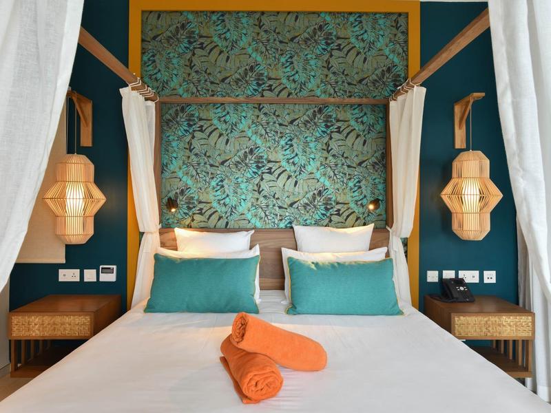 Moderní manželská postel s tyrkysovými polštáři, oranžovými ručníky a dřevěnými lampami v hotelovém pokoji.