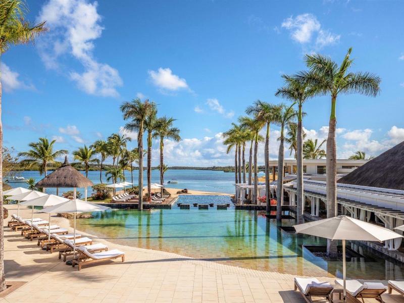 Luxurious pool area with palm trees, sun loungers, and ocean view under a blue sky.