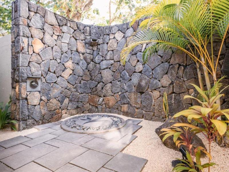 Outdoor shower with natural stone wall and plants in a paved hotel area.
