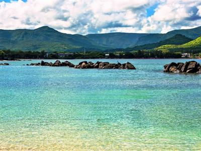 Čistá tyrkysová voda s kameny a zelenými horami pod zataženou oblohou.