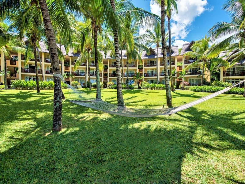 Zelená zahrada s palmami a houpacími sítěmi před resortem pod modrou oblohou.