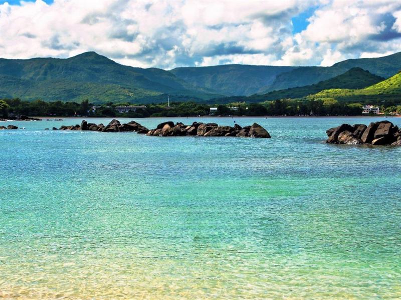 Čistá tyrkysová voda s kameny a zelenými horami pod zataženou oblohou.