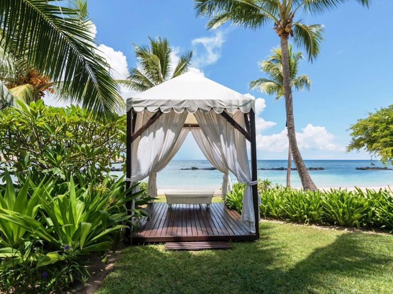 Elegantní pavilon s bílými závěsy na tropické pláži s palmami a výhledem na moře.