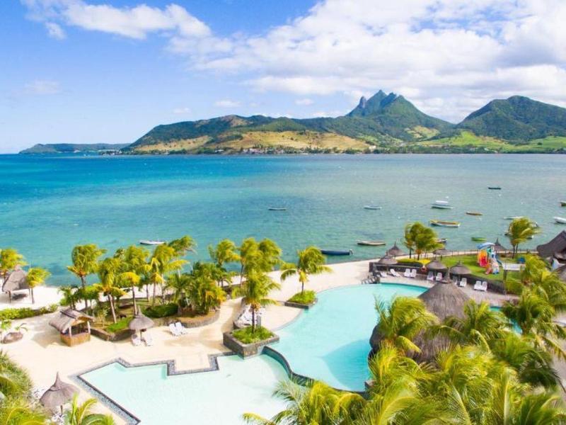 Plage tropicale avec des palmiers, piscine et montagnes en arrière-plan sous un ciel ensoleillé.