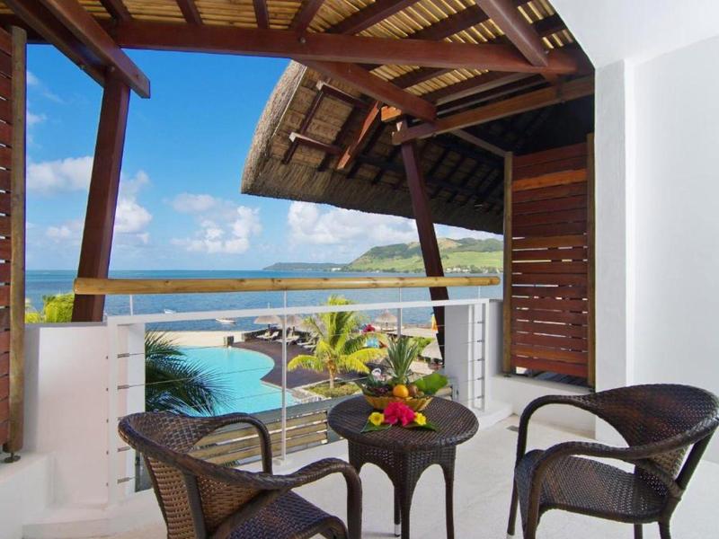 Balcon avec deux chaises et une table donnant sur une piscine, des palmiers et l'océan sous un ciel clair.