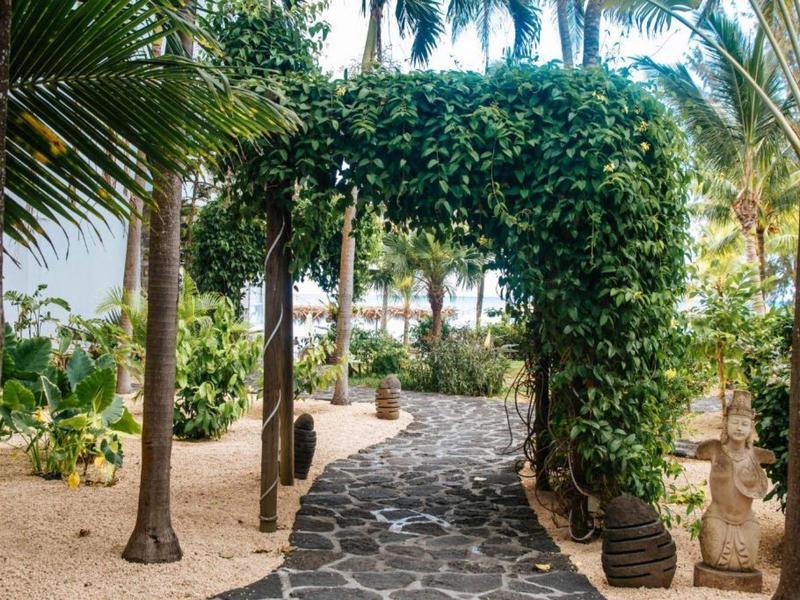 Curved stone pathway with lush green archway amid palm trees in tropical hotel garden.