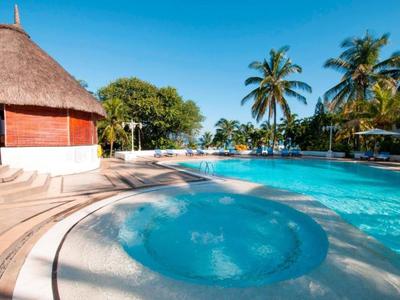 Un'area piscina tropicale con palme, ombrelloni e una vasca idromassaggio sotto un cielo azzurro.