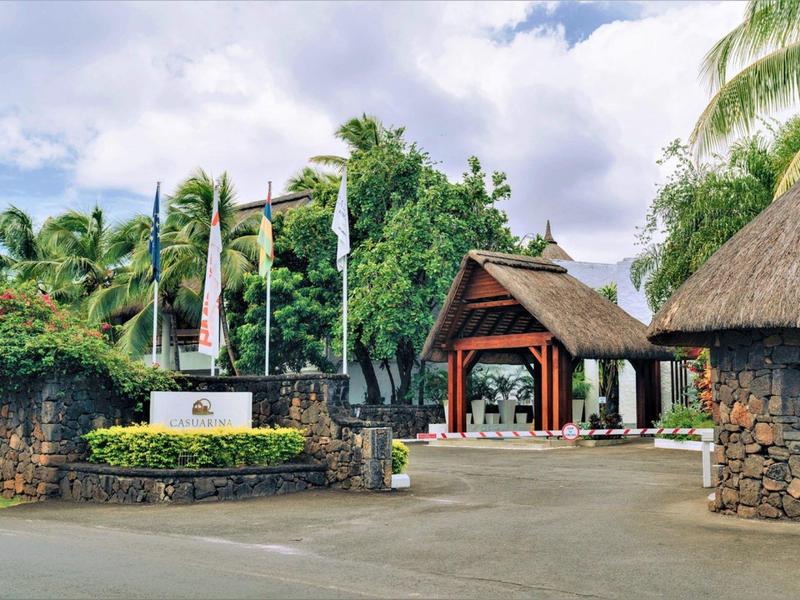 Eingang mit Holzdach und Steinmauern, umgeben von grüner tropischer Vegetation und Palmen.