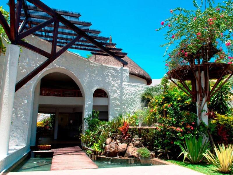 Weißes Haus mit Bogenfenstern, blauer Himmel, grünen Pflanzen und Palmen im Garten.