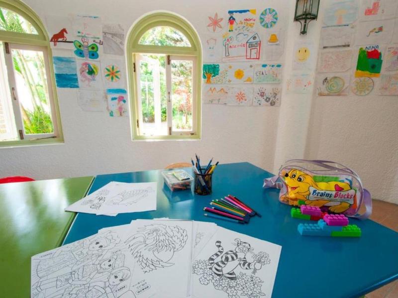 Kindergartenraum mit bunten Malblättern, Stiften, Zeichnungen an weißen Wänden und hellem Fensterlicht.