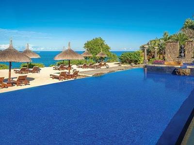 Großer blauer Pool mit Sonnenliegen und Strohhüten am Meer unter blauem Himmel.