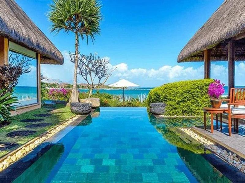 Ein tropischer Pool mit blauem Wasser, umgeben von Palmen, Pflanzen und zwei Strohdachhäusern.